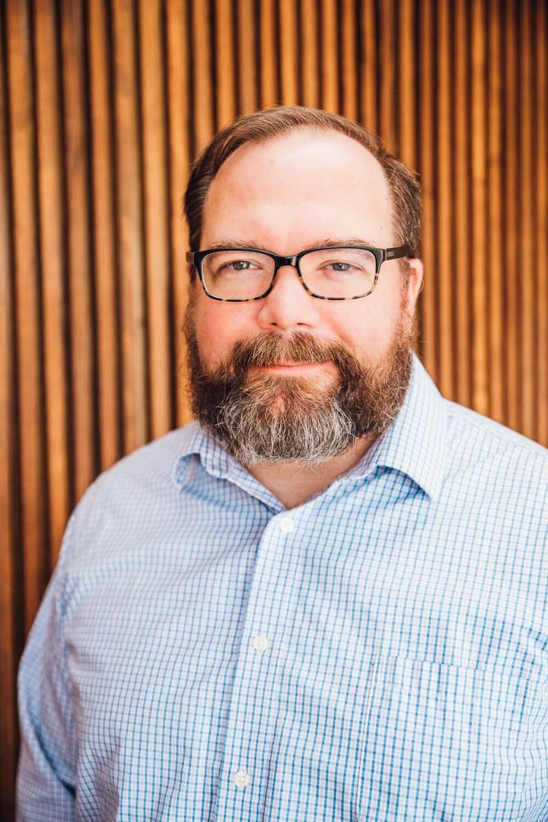 A man with glasses and a beard wearing a blue checkered shirt stands in front of a background with vertical wooden slats.